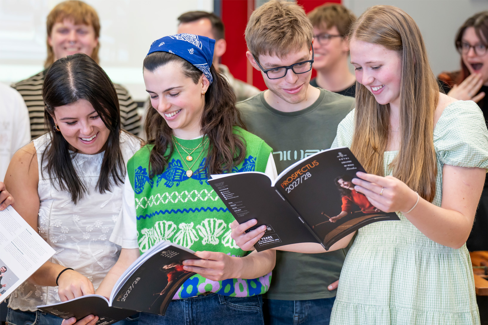 A student wearing a jumper vest, reading a booklet to people next to her, smiling.